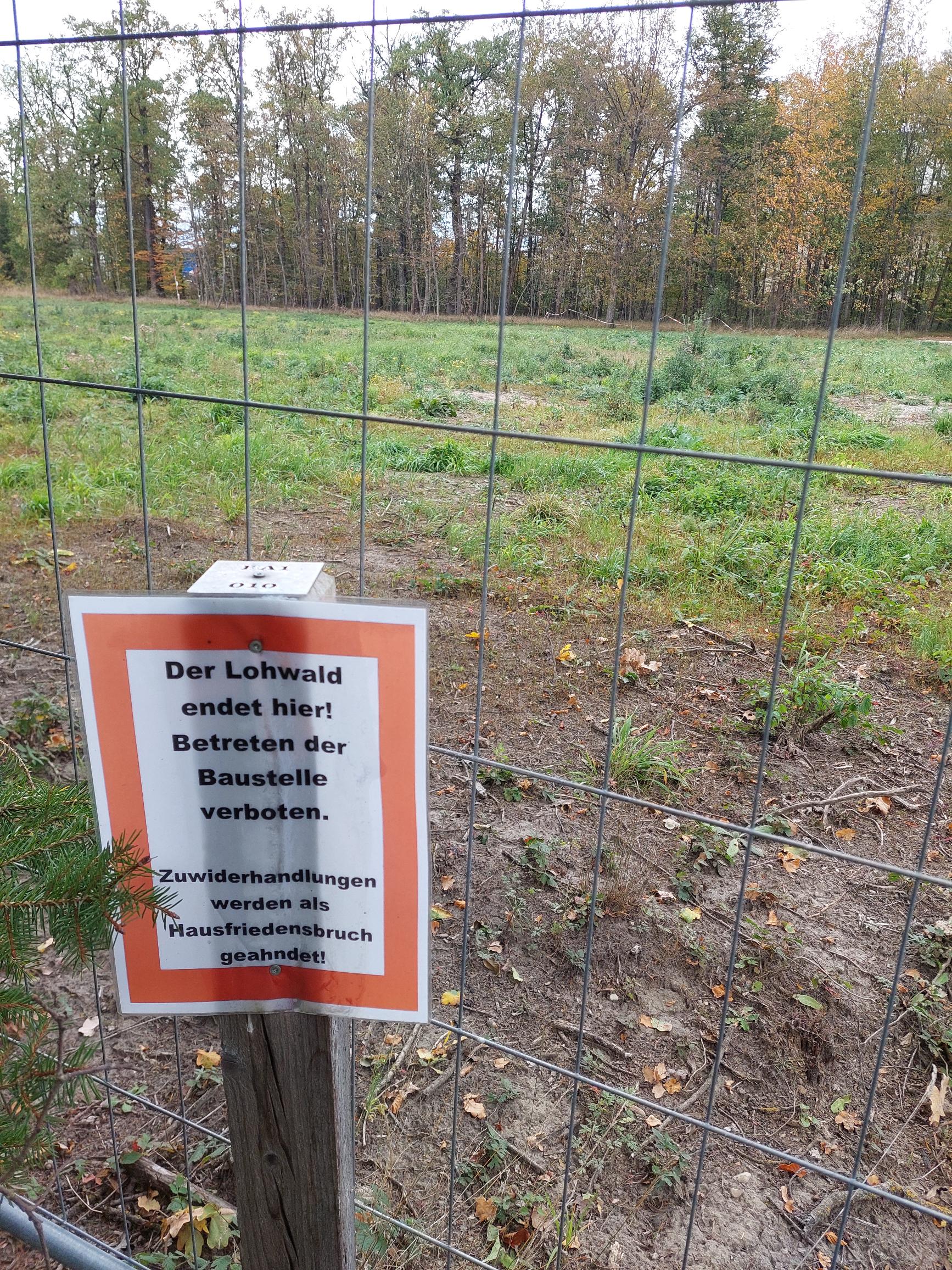 Ein Bauzaun versperrt den Weg auf eine Grasfläche
       mit großen grasfreien Stellen.
       Vor dem Zaun steht ein Schild mit der Aufschrift
       „Der Lohwald endet hier! Betretten der Baustelle verboten.
       Zuwiderhandlungen werden als Hausfriedensbruch geahndet!“.